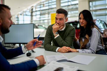 A close-up of a casually dressed couple sitting at a table, happily listening to the car salesman...