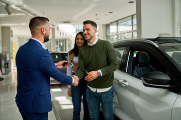 A close-up of a casually dressed couple standing next to a white car, facing us. The salesman in a...