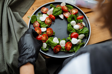 A person in stylish black gloves is skillfully preparing a vibrant salad, artfully arranging fresh...