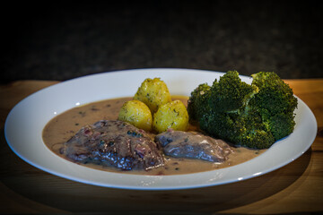 ready to eat, roasted game meat from chamois with pepper sauce, broccoli and potatoes, served on a white plate and a rustic wood board
