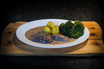 ready to eat, roasted game meat from chamois with pepper sauce, broccoli and potatoes, served on a white plate and a rustic wood board