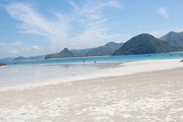 A tranquil beach stretches along the shoreline with soft, white sand. Gentle waves lap at the coast while distant green hills rise against a bright blue sky, creating a peaceful atmosphere.