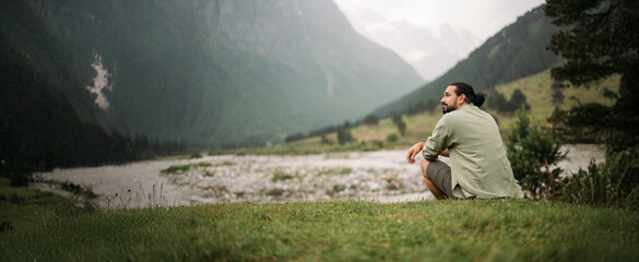Portrait of a male tourist sitting on the riverbank against the background of misty mountains.  A...