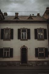 an old, weathered building with a faded white facade, closed shutters, and a dark entrance. The gloomy sky enhances its rustic charm