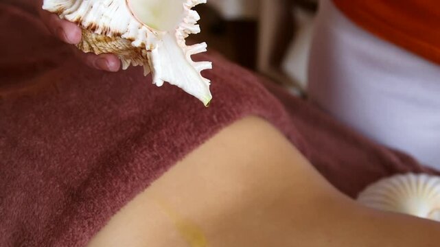 Masseuse Pours Oil From Beautiful Shell On Naked Woman's Back Before Massage. Different Types of Massage Cremes, Lotions, Gels, and Oils are Used to Lubricate and Moisturize the Skin.