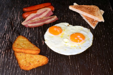 A delicious breakfast meal consists of fried eggs, bread toast, bacon and sausages isolated on a wooden surface.