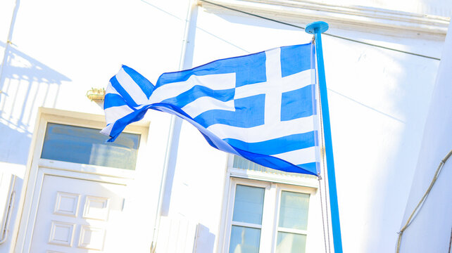 Bandeira da Gr&eacute;cia tremulando em uma viela de Mykonos, Gr&eacute;cia, com suas t&iacute;picas casas brancas e ruas estreitas, capturando a ess&ecirc;ncia da cultura e arquitetura grega