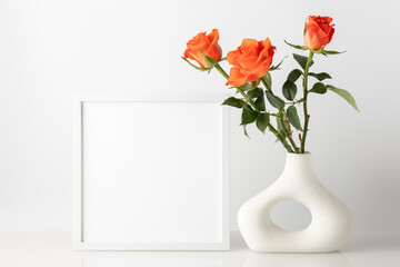Bouquet of natural coral orange rose flowers in modern donut shape vase and empty photo frame on glass table against white wall. Home interior decoration.