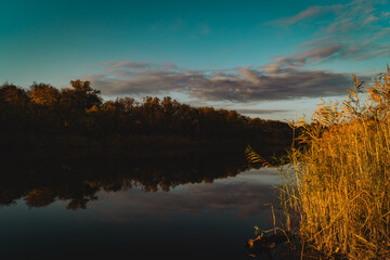 a serene landscape featuring a calm river at sunset. On the left side of the image, tall grass and reeds with golden hues line the edge of the water, indicating the warm light of the setting sun