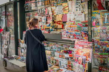 Hesitant Woman Looking at Magazines and Newspapers in a Street Kiosk, Thinking Which One to Buy....