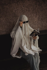 A figure draped in a white sheet resembling a ghost sits on a bench, donning sunglasses and reading a book. The backdrop features textured walls, lending a mysterious vibe.