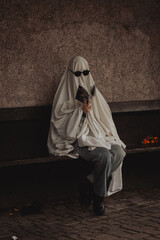 A figure draped in a white sheet resembling a ghost sits on a bench, donning sunglasses and reading a book. The backdrop features textured walls, lending a mysterious vibe.