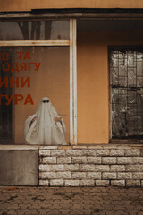 Naklejka premium A ghostly figure draped in white stands behind a shop window, wearing dark sunglasses. The window features bold orange text, while the exterior shows a weathered facade
