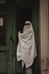 A figure in a white sheet resembling a ghost stands by a green door, wearing oversized sunglasses. The atmosphere feels eerie yet playful, evoking a Halloween vibe.