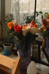A vibrant bouquet of crocheted flowers in a purple vase sits on a wooden table beside a mirror. Natural light filters through sheer curtains, illuminating green plants and pottery.