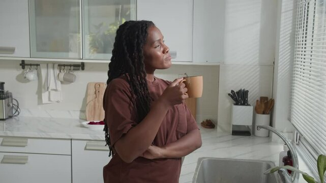 Black young woman drinking coffee and daydreaming while standing near window in kitchen having leisurely morning