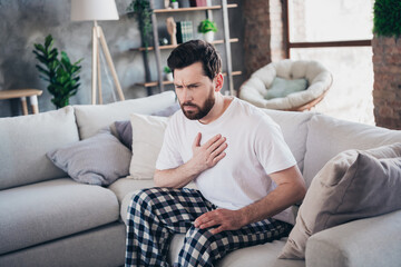 Photo of young man wear pajama sit on sofa arm touch chest heart attack pain hurt living room indoors