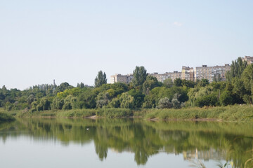 Fototapeta premium A serene river reflects a blue sky and lush greenery. In the distance, modern apartment buildings rise, blending urban life with nature, creating a tranquil scene.