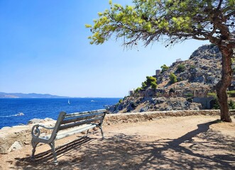 Photo of an idyllic view from Hydra, Greece, featuring a shaded bench under a pine tree overlooking the azure Aegean Sea, distant sailboats, and the island's iconic rocky hills.