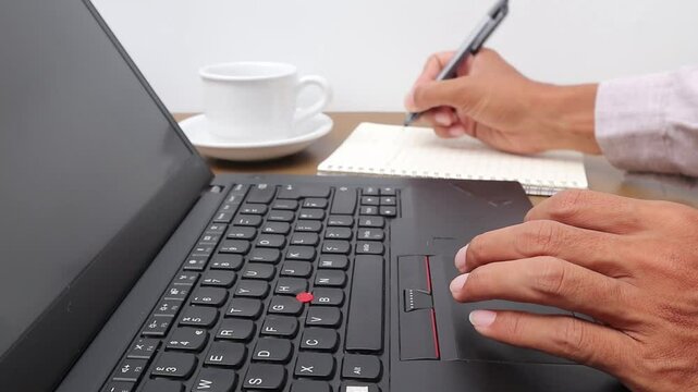Man's hand writing in notebook while looking for references on laptop
