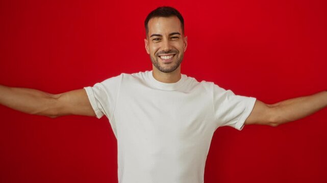 Young hispanic man posing over isolated red background showing cheerful expressions and strong arm muscles while wearing a plain white t-shirt