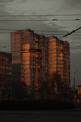 a tall, gray apartment building at dusk, illuminated by warm light reflecting off its windows, amidst a moody sky filled with clouds and power lines.