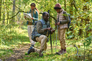 African American father kneeling next to son and teaching him how to use compass while his older kid exploring map in background