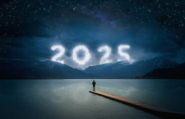 New year 2025 at night, man standing on a wooden jetty on a lake and looking to the cloudy numbers in the dark sky with stars over mountains, copy space