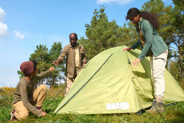 Low angle shot of African American boy sitting on grass and helping his parent and sister with pitching tent in forest while they camping together
