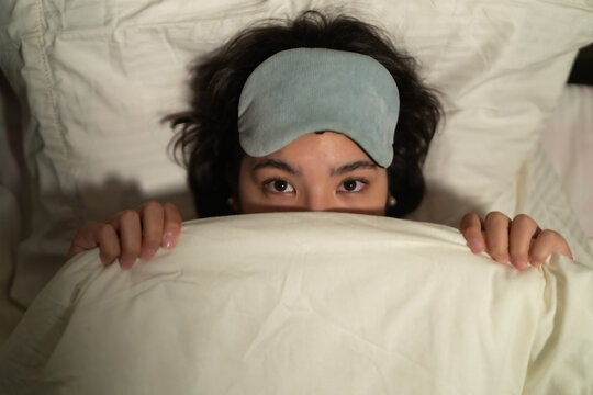 Young woman hiding under blanket suffering from insomnia wearing sleep mask