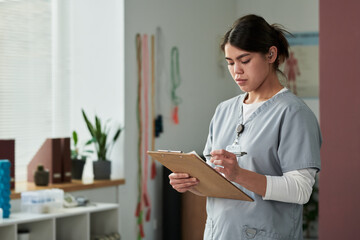 Medical professional writing on clipboard in organized office with medical tools visible in...