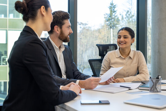 Job interview scene with young woman engaged with two professionals in bright office setting. People are seated at table with documents, discussing opportunities in modern workspace.