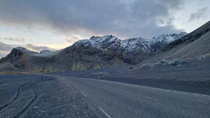 Naklejka premium Snowy mountain cliffs across long road in icelandic nature landscape, highway in wintry weather. Peaks covered in snow alongside lonely road within beautiful nordic valley.