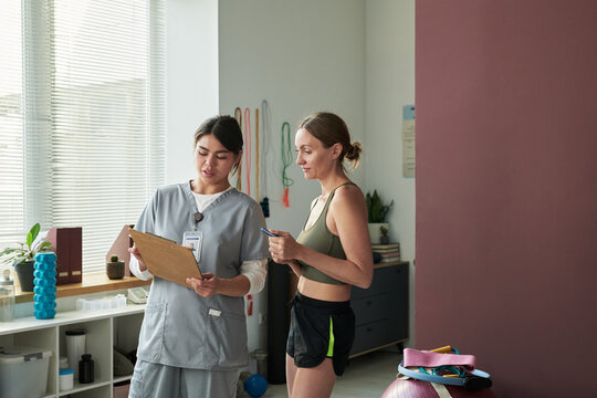 Woman in workout attire conversing with fitness professional holding clipboard at gym. Sunlight streaming through windows and exercise equipment seen in background