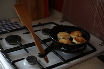 Golden-brown cheese pancakes frying in a black pan on a gas stove. A retro-styled teapot is visible in the background, creating a cozy and homely atmosphere