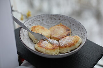 A plate of golden-brown syrniki (cottage cheese pancakes) dusted with powdered sugar, served with a fork on a patterned ceramic dish