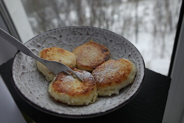 A plate of golden-brown syrniki (cottage cheese pancakes) dusted with powdered sugar, served with a fork on a patterned ceramic dish