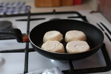 Raw syrniki (traditional cottage cheese pancakes) frying on a gas stove in a black non-stick pan....