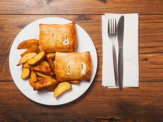 Dinner meal with golden wedges and two crispy fish and cheese pie on white plate and wooden table. Fine food with fried potatoes and staffed pastry. Simple basic dish.