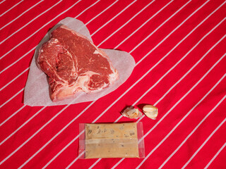 Fresh uncooked T bone steak on heart shaped cooking paper and garlic cloves and pouch with sauce on...