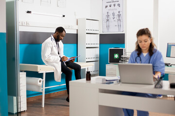 Caucasian nurse working on laptop while african american doctor sits on hospital bed reviewing medical information on clipboard. Diverse healthcare staff in clinic office preparing to treat patients.