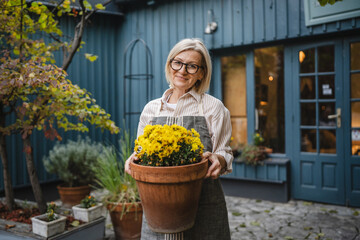 Mature woman waitress hold potted yellow flowers outside rustic shop