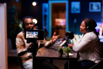 Black man creates vlogs with a digital camera and produces podcasts with professional audio gear. He interviews guests in his home studio, filming with a tripod.