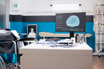 Wheelchair user doctors table with desktop pc showing brain imaging in vacant clinic room, ideal for health assessments. Tools, files, and technology are arranged for effective patient care.