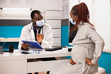 African american doctor and pregnant woman engage in prenatal consultation in medical office. Physician speaks on maternal health while expectant mother listens with hands resting on pregnancy belly.