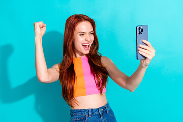 Joyful young redhead girl in vibrant summer outfit celebrates success through her phone against a...