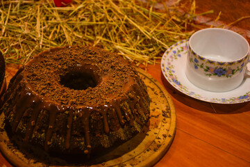 A cozy scene of a slice of rich chocolate cake next to a steaming cup of tea. The soft, warm lighting emphasizes the indulgence and comfort of the moment.