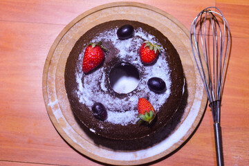 A close-up shot of a rich, moist chocolate cake with glossy chocolate frosting and a piece of fresh fruit as decoration. The cake is placed on a rustic wooden table with soft lighting.