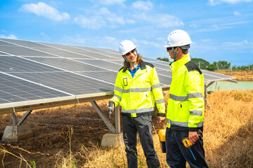 Two engineers wearing safety helmets and reflective jackets inspect a solar panel installation. One uses a drill tool, while the other checks the panel surface under a bright, sunny sky.