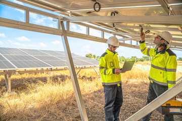 Two technicians in high visibility jackets and helmets are inspecting the underside of solar panels at a solar farm. One holds a drill, while the other uses a laptop for analysis and reporting.